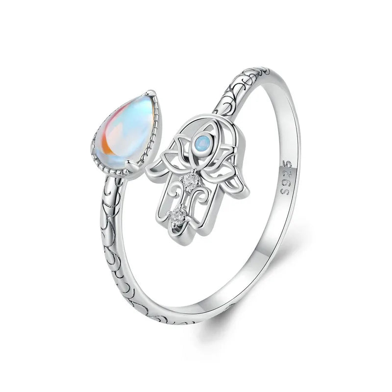 A close-up image of a silver adjustable ring featuring a Hamsa Hand design with intricate details and a blue stone centerpiece. Opposite the Hamsa Hand is a teardrop-shaped opal stone, displaying an iridescent glow. The band includes engraved patterns, adding an elegant and meaningful touch to the jewelry.